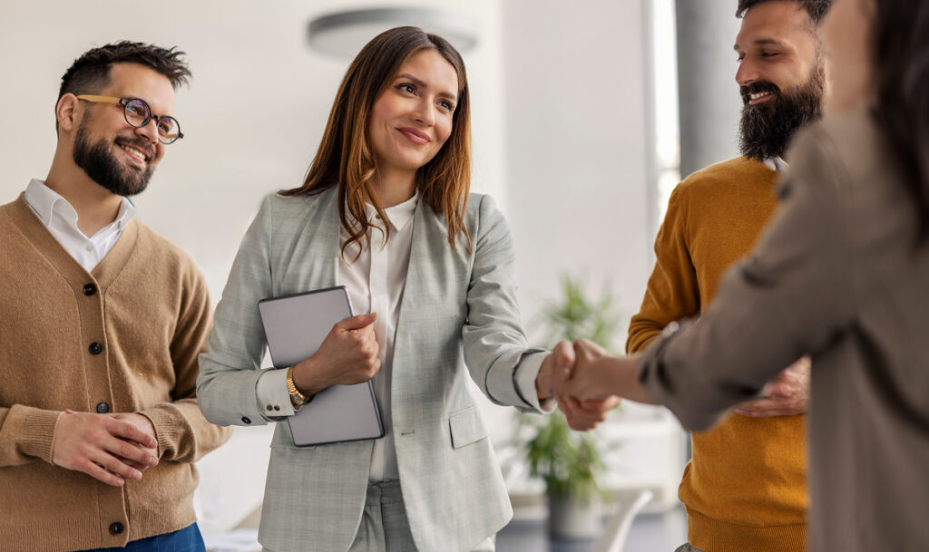 Female chief shaking hands with business partner while standing with her team of experts at office.